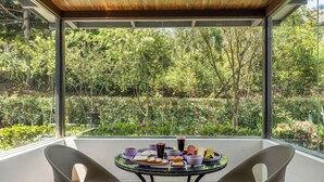 A balcony with a table set for breakfast, surrounded by glass walls offering a view of a lush garden. - Kalpavriksha Villa By Indostay (Kotagiri)