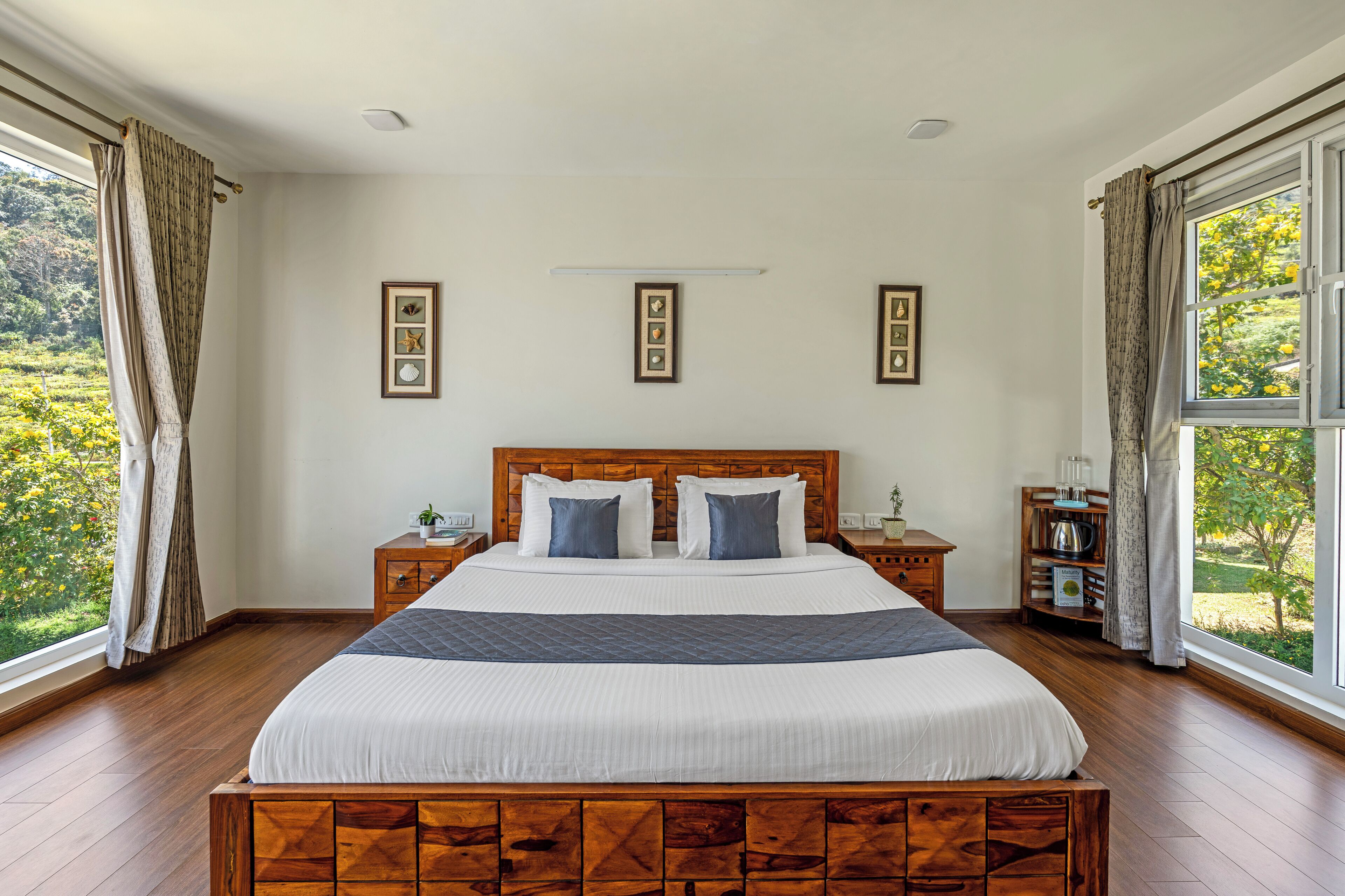 A large bed with a wooden headboard, two bedside tables, and a nightstand with a lamp. There are framed pictures on the wall and a window with curtains.