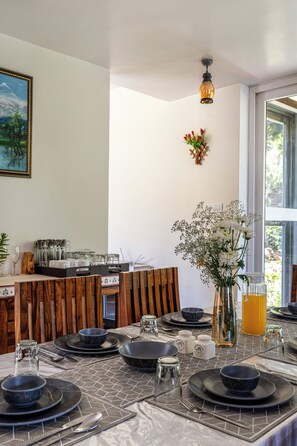 Un comedor con una mesa dispuesta para cuatro personas, con platos, cuencos y cubiertos negros. Se aprecia un jarrón con flores blancas sobre la mesa, y una silla de madera al fondo.