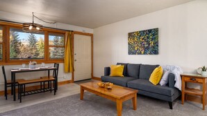Un salon comprenant un canapé, une table basse, une table à manger et des chaises. Il y a un tableau au mur et une fenêtre avec des rideaux.
