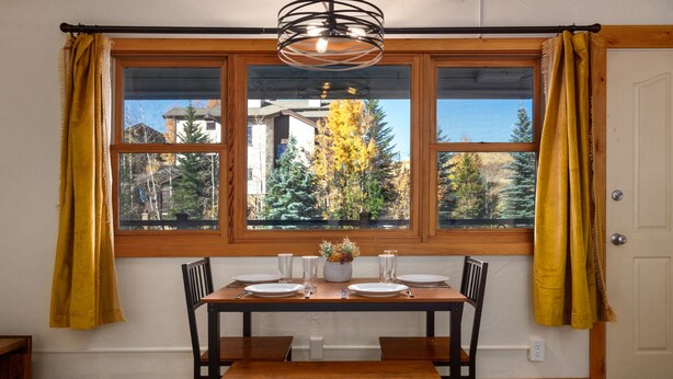 Un coin repas avec une table et des chaises en bois, une fenêtre avec des rideaux et une vue sur les arbres et les bâtiments à l’extérieur.
