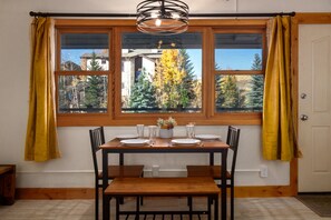 A dining area with a wooden table and chairs, a window with curtains, and a view of trees and buildings outside. - Ski In/Out, Hot Tub, Walk to Shops, Free Bus +EZCancel (Steamboat Springs)