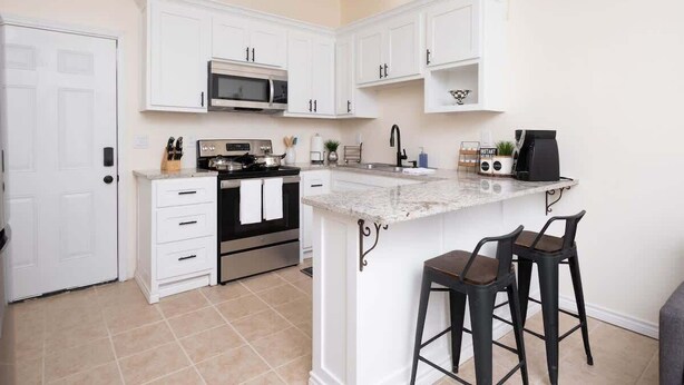 A kitchen with white cabinets, a microwave, a stove, a sink, and two bar stools. - 1BR Sleeps 4 | AI Data Ctr, Hospitals, Corp Stays (Amarillo)