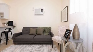 A living room with a grey sofa, a small round table with a sign, a chair, and a white radiator on the wall. - 1BR Sleeps 4 | AI Data Ctr, Hospitals, Corp Stays (Amarillo)