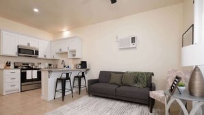 A modern kitchen with white cabinets, a microwave, and a stove. There is a gray sofa with a green throw blanket and a small table with a lamp and decorative items. - 1BR Sleeps 4 | AI Data Ctr, Hospitals, Corp Stays (Amarillo)