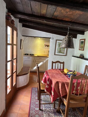 Een eetgedeelte met een houten tafel, stoelen en een rode tafelkleed. Achtergrond is een keuken met een houten plafond en een hangende lamp.