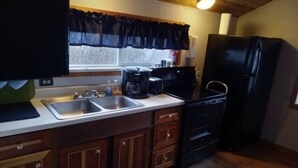 A compact kitchen with wooden cabinets, a black refrigerator, a coffee maker, and a window with a dark curtain. - Best View of the Lake in Harrison (Harrison)