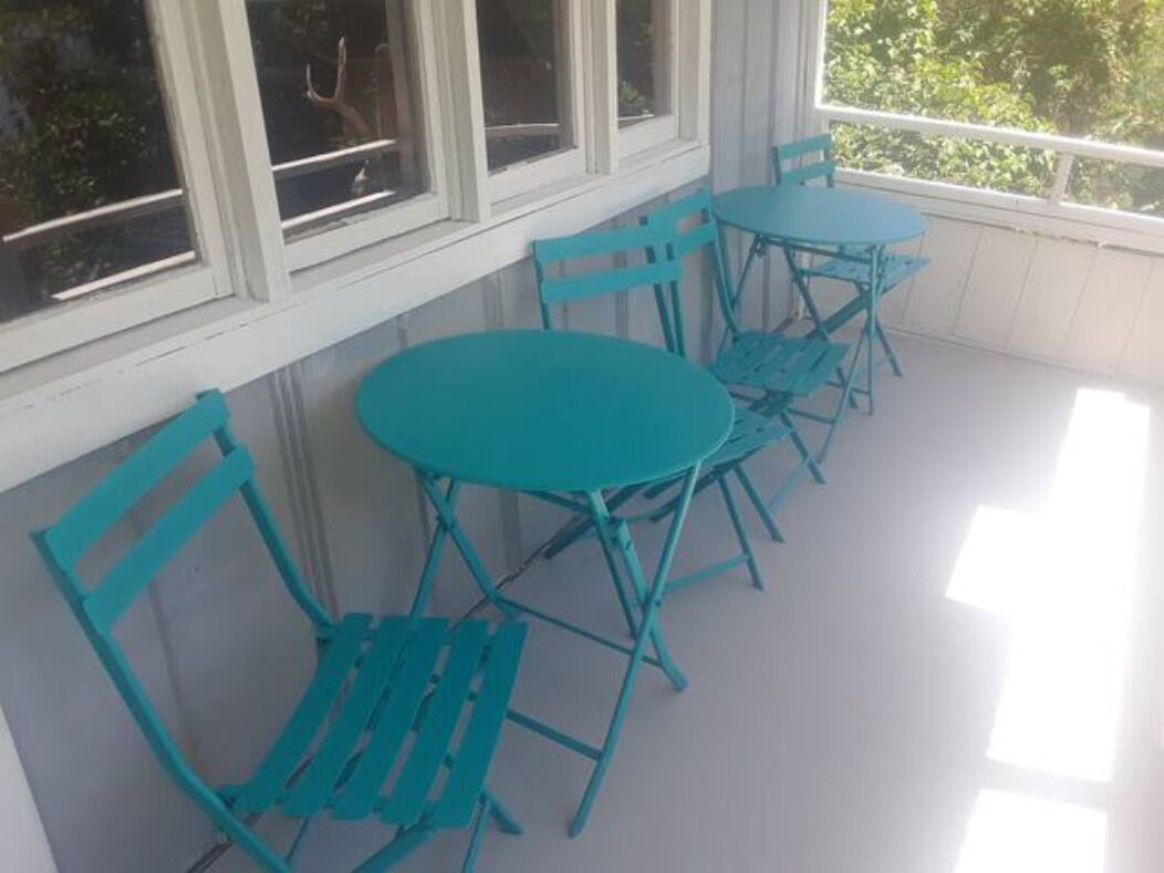 Un'area salotto all'aperto coperta, con sedie e tavoli in metallo color turchese, pareti bianche e finestre che si affacciano sul verde.