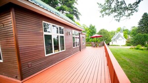 A wooden deck with a red railing, a white building with a green roof, and a red umbrella in the background. - Entire Home located in historical district (Bayfield)