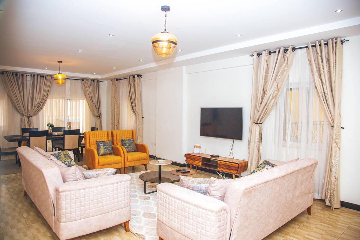 Sala de estar com dois sofás, uma mesa de centro e uma televisão. Ao fundo, uma mesa de jantar com cadeiras.