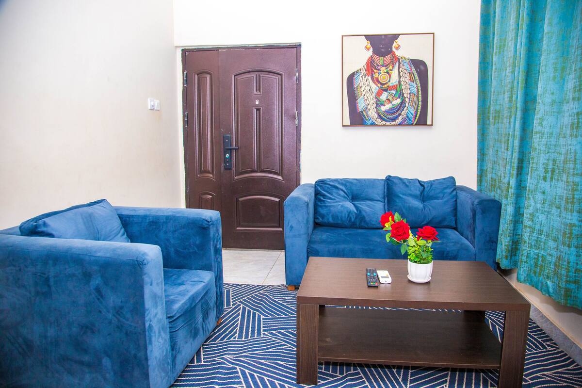 Quarto com sofá azul, mesa de centro de madeira com um vaso de flores vermelhas e uma porta com uma pintura decorativa acima.