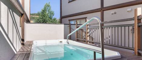 A wooden deck with a glass-enclosed hot tub, a metal railing, and a building with a window and a door.