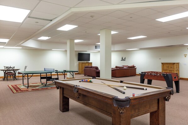Une table de billard avec des billes, une table de baby-foot, une table de ping-pong et un téléviseur dans une pièce dotée d’un plafond haut et d’un sol recouvert de moquette.
