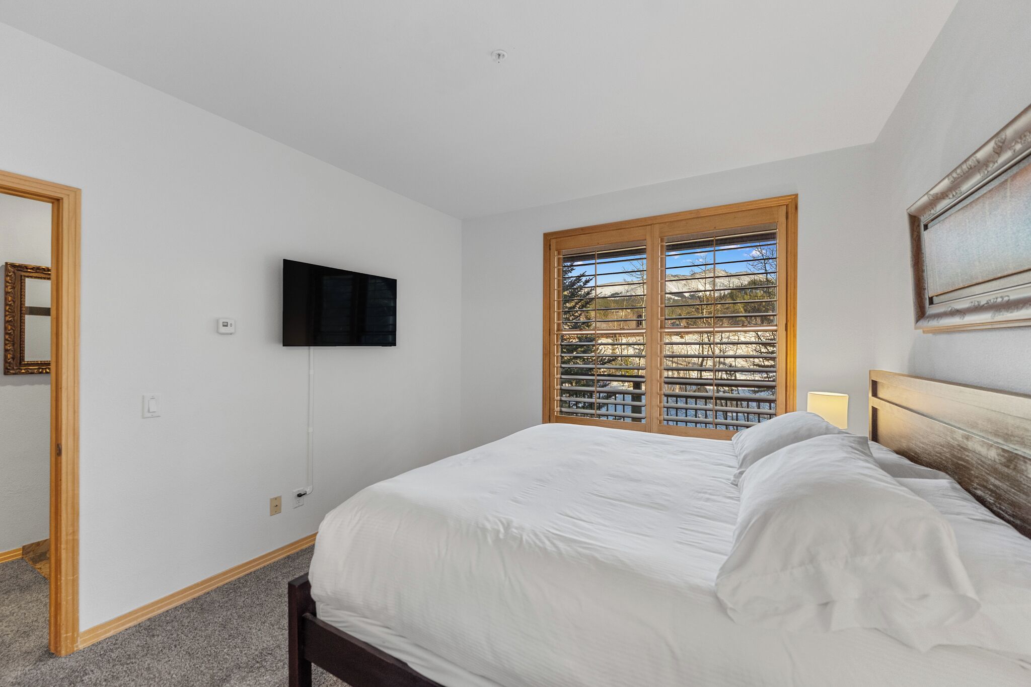 Ein Schlafzimmer mit einem Bett, einem an der Wand montierten Fernseher, einem Fenster mit Jalousien und einer Tür.