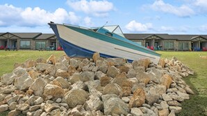 Un bote reposando sobre un conjunto de rocas frente a una hilera de edificios, con cielo despejado.