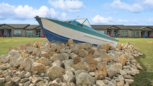 Un bote reposa sobre un montón de rocas frente a una hilera de edificios con ventanales amplios.