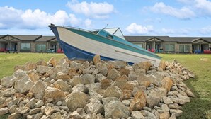 A boat resting on rocks in front of a row of buildings with a grassy field and a clear sky. - MotorBoat Inn Suite 14 (Graford)