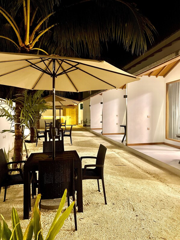 An outdoor dining area with a table and chairs under a large umbrella, surrounded by palm trees and a building with a white exterior. - Vilu Residence (Maamigili)