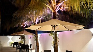 An outdoor seating area with tables and chairs under a large umbrella, surrounded by palm trees and a white wall. - Vilu Residence (Maamigili)