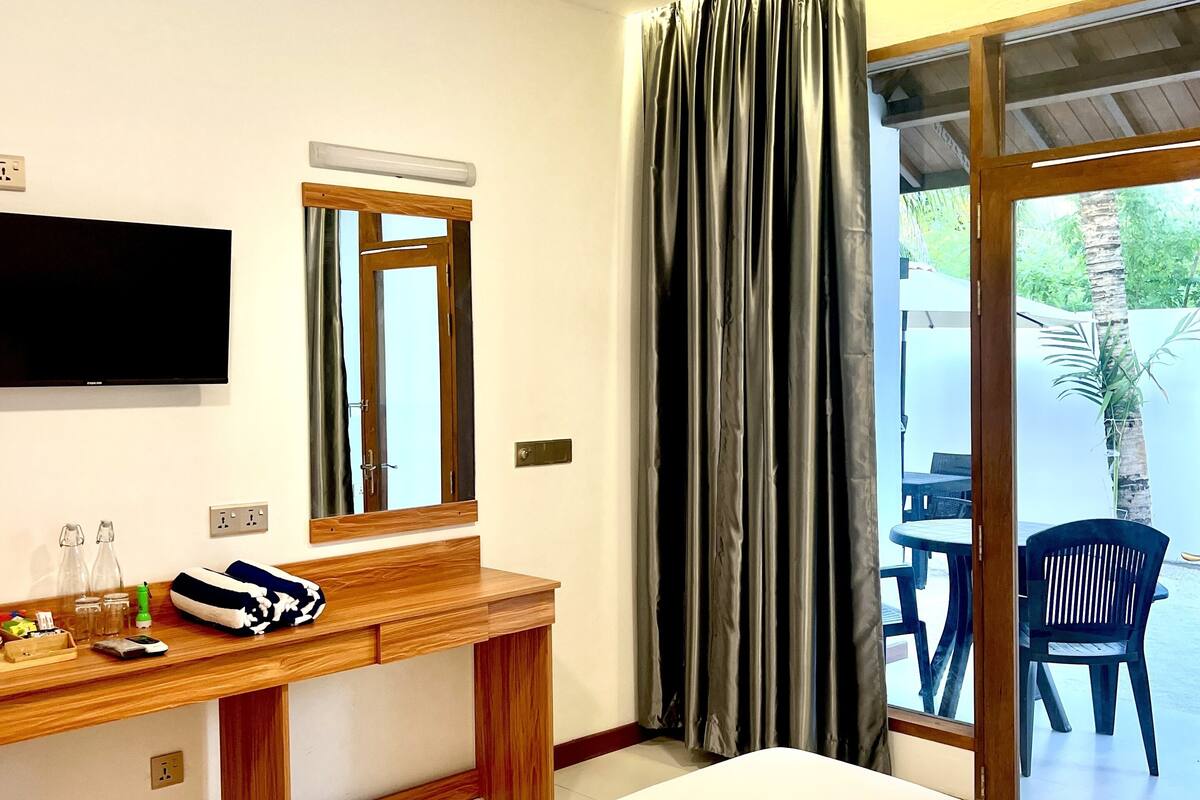 A hotel room with a bed, a wooden desk, a flat-screen TV, a ceiling fan, and a view of the outdoors through a sliding glass door.