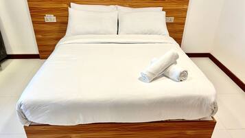 A neatly made bed with white linens and a wooden headboard, a rolled white towel on the bed, and a power outlet on the wall.
