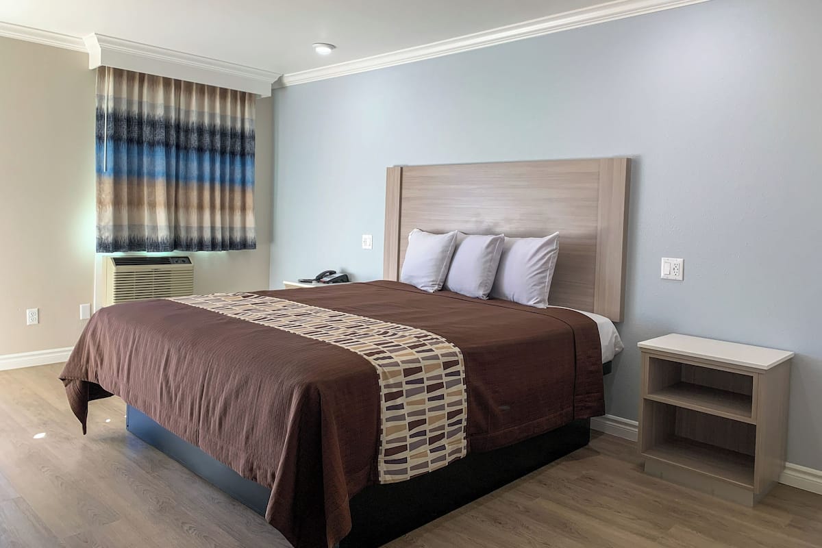 A bedroom with a bed, a bedside table, a nightstand, a window with curtains, an air conditioning unit, and a wall socket.