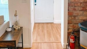 Une pièce avec un plancher en bois, une porte blanche, une petite table avec une lampe et un mur en briques.