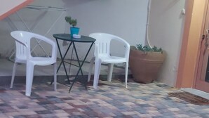Un petit espace extérieur aménagé avec deux chaises en plastique blanches et une table, un pot de fleurs et un sol carrelé aux couleurs vives.