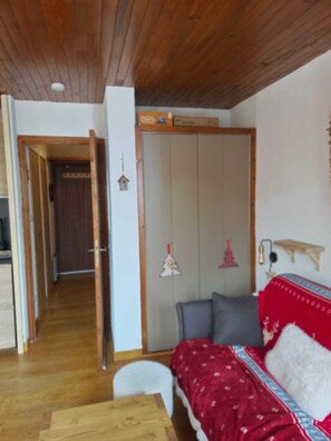 Una habitación con techo de madera, una cama con manta roja, una mesita pequeña y un armario.