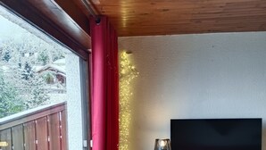 A room with a wooden ceiling, a large window with red curtains, a TV on a wooden cabinet, and a small table with a lamp.