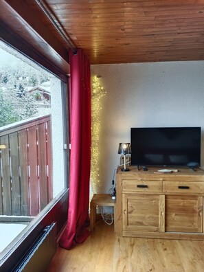 Una habitación con techo de madera, un ventanal con cortinas rojas, un televisor sobre un mueble de madera y una mesita con una lámpara.