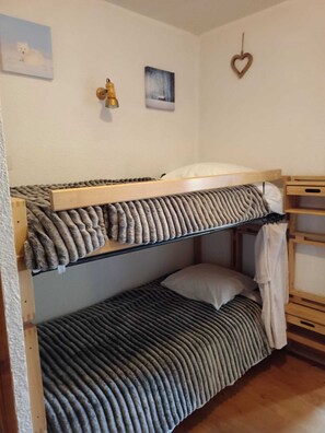A bunk bed with a wooden frame and a grey striped mattress. There is a small shelf with a white pillow and a heart-shaped decoration on the wall.