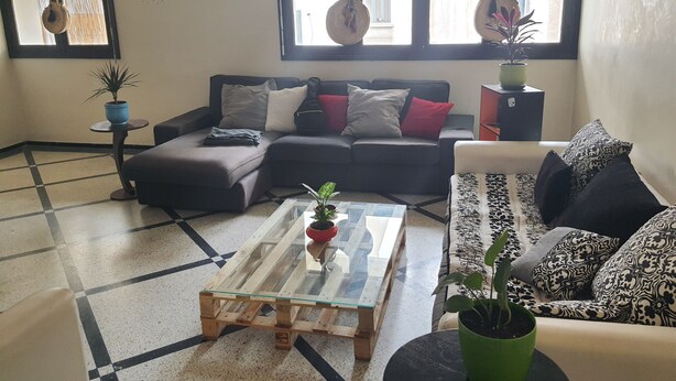 Un salon avec un canapé d’angle, une table basse en verre et un fauteuil à motifs. On y trouve deux plantes en pot et une petite table d’appoint.