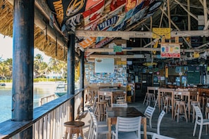 An outdoor dining area with wooden tables and chairs, a thatched roof, and various signs and posters.