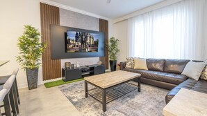 En moderne stue med et fladskærms-tv på væggen, en brun lædersofa, et træsofabord og to krukker med planter.