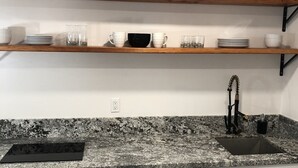 A kitchen with white cabinets, a granite countertop, a black sink, and wooden shelves above holding dishes and glasses.