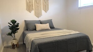 A bedroom with a bed, grey bedding, a plant, and macramé wall hangings.