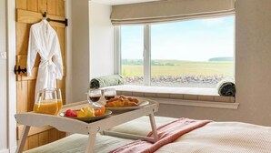 A bedroom with a bed, a breakfast tray with croissants and fruit, a window with a view of the countryside, and a robe hanging on a wooden door. - South Riccalton Farm Cottage (Jedburgh)
