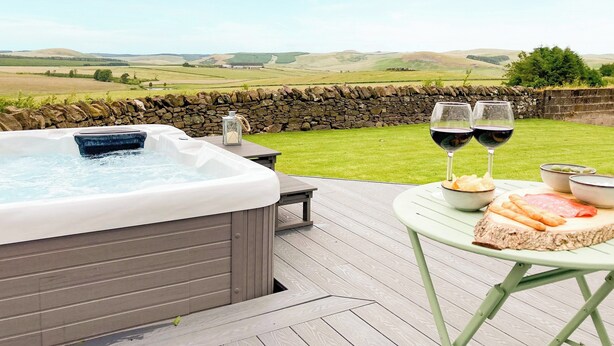 A hot tub on a wooden deck with a stone wall and a scenic view of rolling hills. - South Riccalton Farm Cottage (Jedburgh)