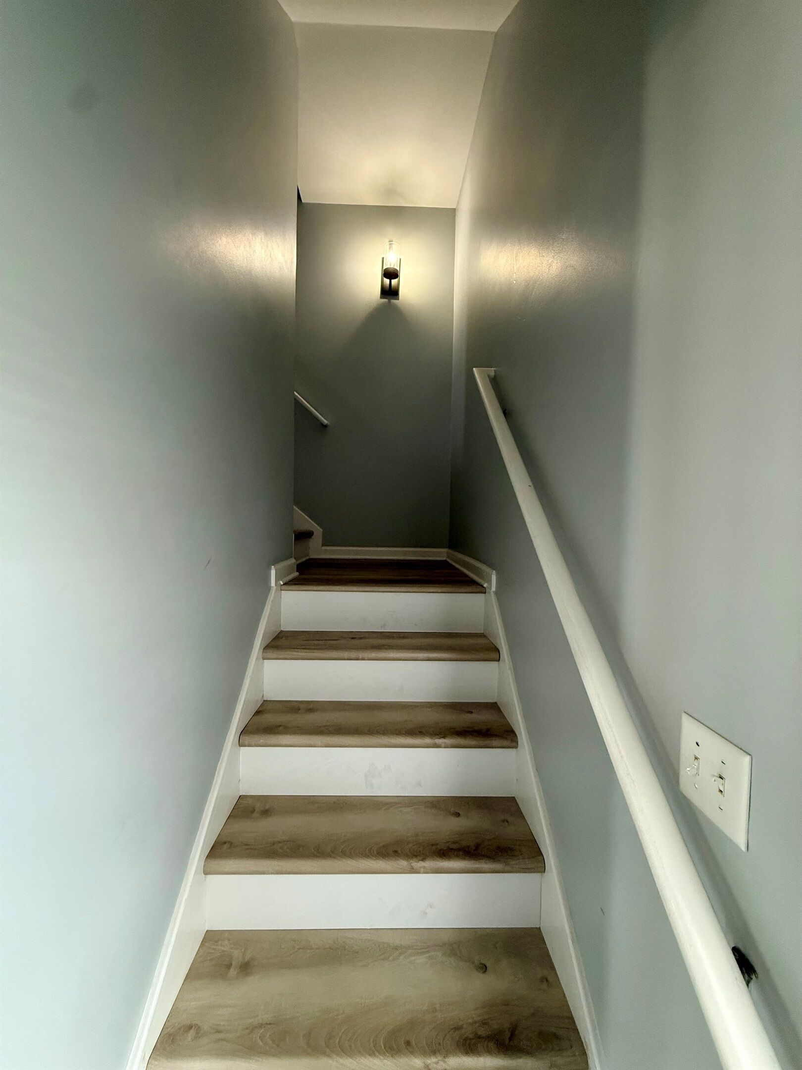 Una escalera de madera con barandillas blancas que lleva a un rellano con una lámpara fijada a la pared.