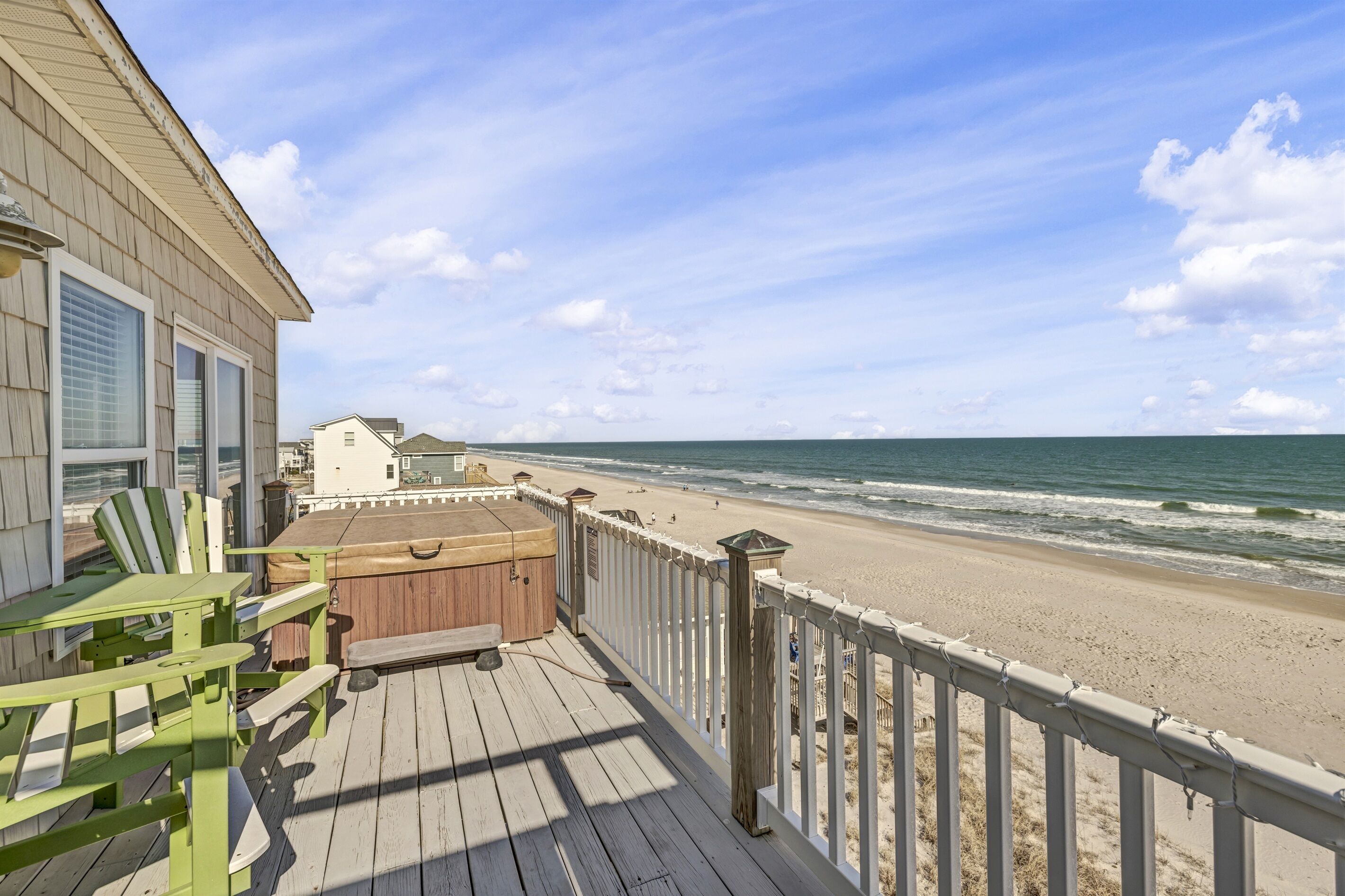 A wooden deck with green chairs and a hot tub overlooking a beach.