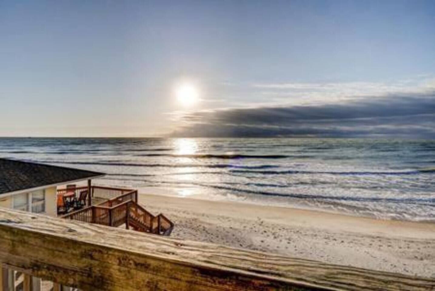 Une propriété située en bord de mer, dotée d’une terrasse en bois offrant une vue sur l’océan, sous un ciel dégagé, et où l’on peut admirer le soleil se coucher sur l’eau.