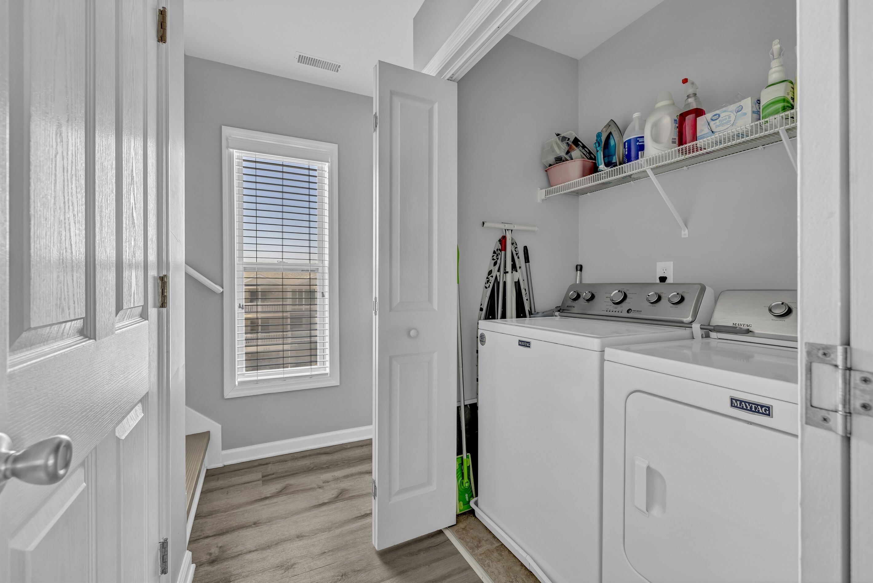 A laundry room with a Maytag washer and dryer, a window with blinds, and a shelf with cleaning supplies.