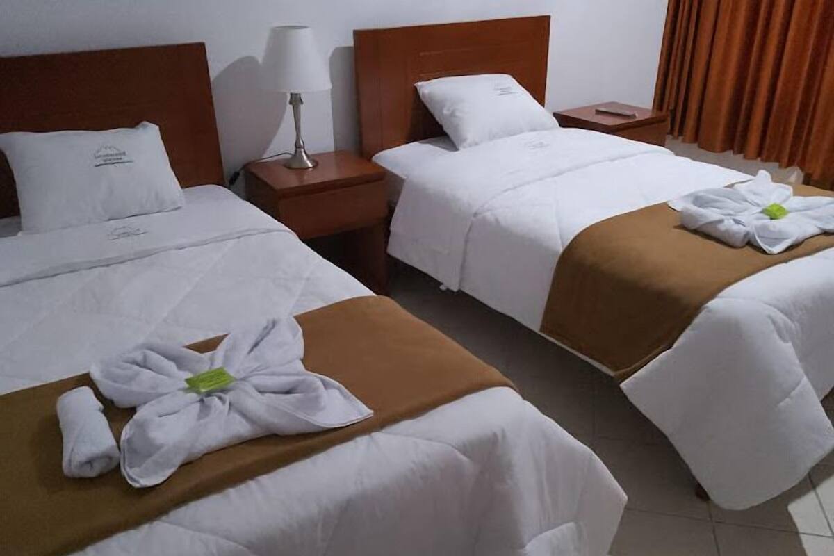 A hotel room with two beds, each with white bedding and brown headboards. There is a nightstand with a lamp between the beds.