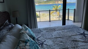 Un dormitorio con una cama grande, una mesita de noche, una lámpara y vistas a una playa y al océano a través de una puerta corrediza de vidrio.