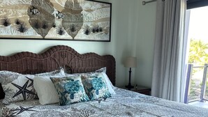 Un dormitorio con una cama grande, cabecera, una obra de arte en la pared, una mesita de noche con lámpara y una ventana con cortinas.