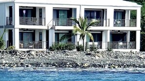 Un edificio blanco de varios pisos con balcones, ubicado en una costa rocosa con una palmera frente a él.
