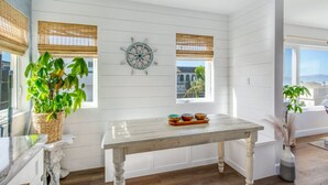 Una habitación con paredes de paneles blancos, una mesa de madera con cuencos decorativos, un adorno de timón de barco y una planta grande en maceta.