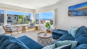 Un salón amplio con un gran ventanal, un conjunto de sofá azul, una mesa de centro redonda y un cuadro con una playa.