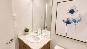 A modern bathroom with a double sink, wooden cabinets, a toilet, a mirror, and a wall-mounted light fixture. - Perfect Studio Downtown Sidney (Victoria)